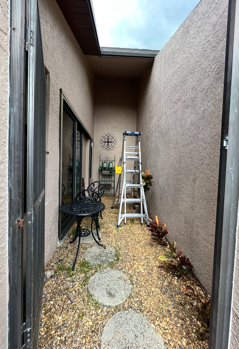 A narrow, gravel-floored patio with a table, chair, and ladder, flanked by walls and accessible by a gate.
