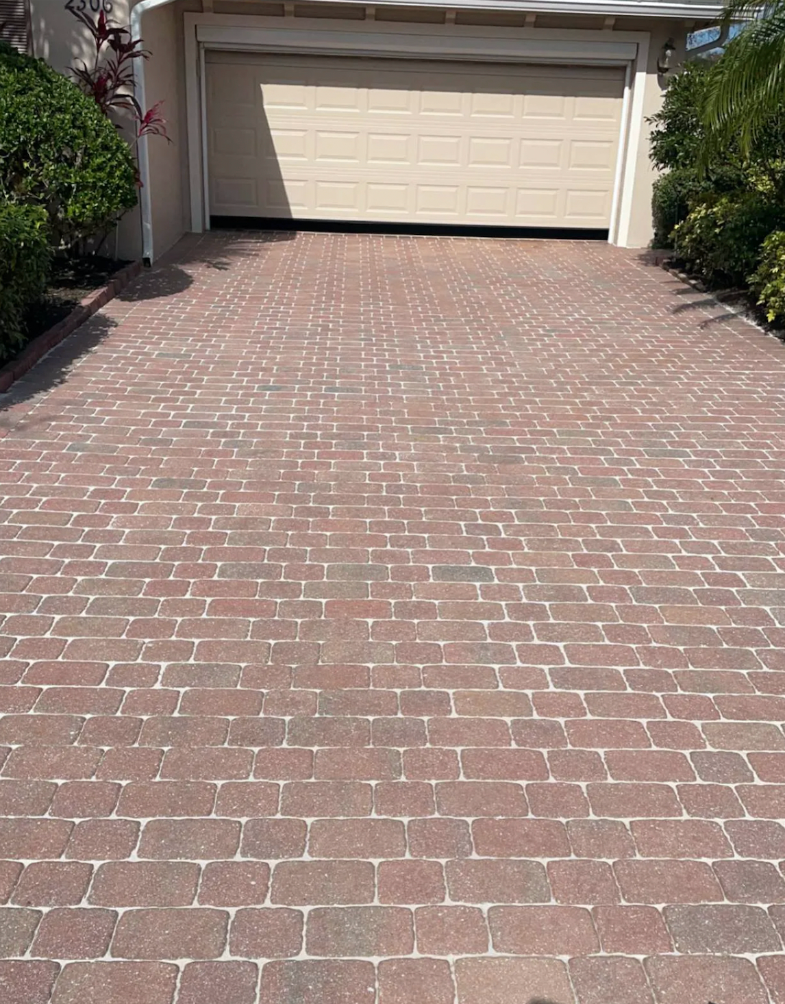 Brick driveway leading to a closed garage door, flanked by green bushes.