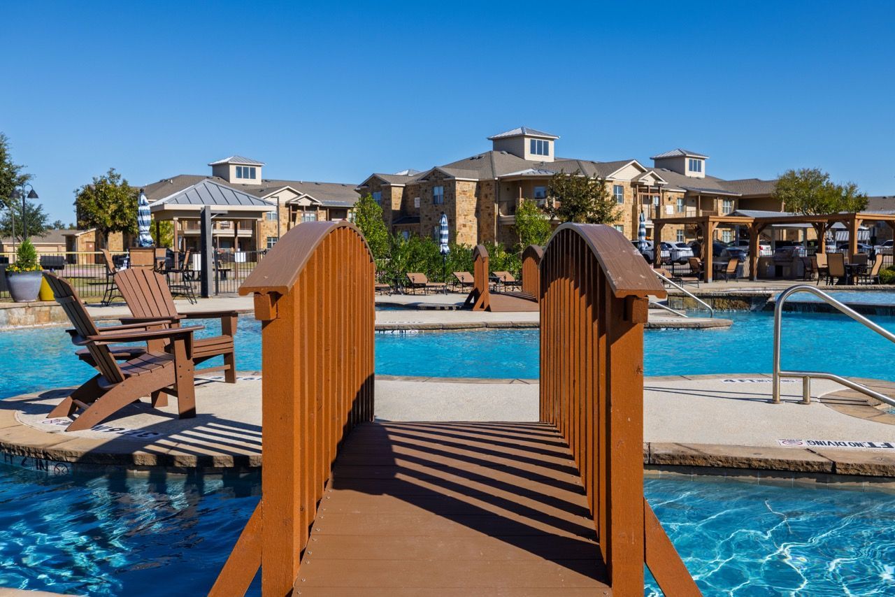 Brown wooden bridge over a bright blue pool with lounge chairs and buildings in the background.
