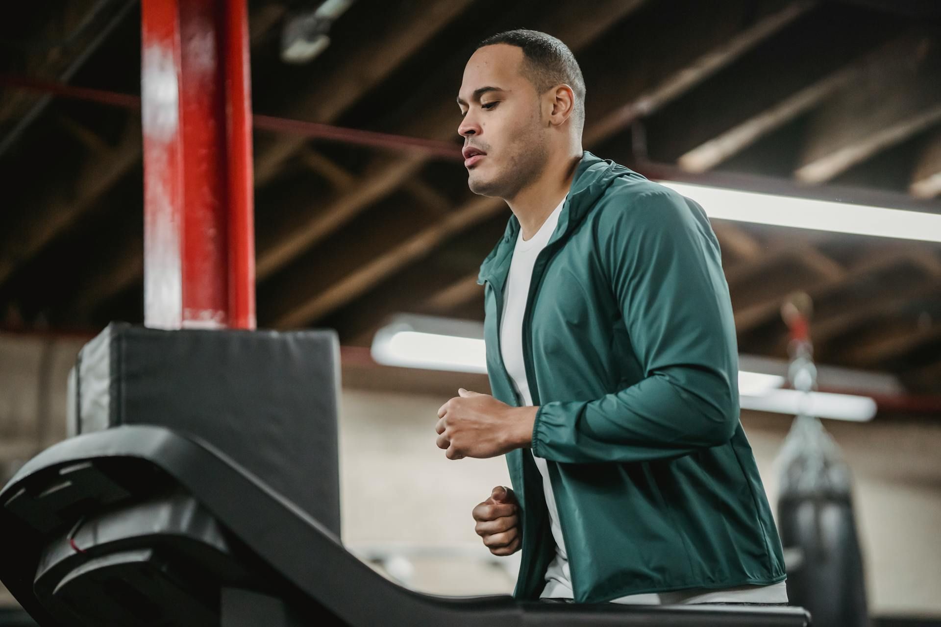 Man in teal jacket runs on a treadmill in a gym with red support beams.