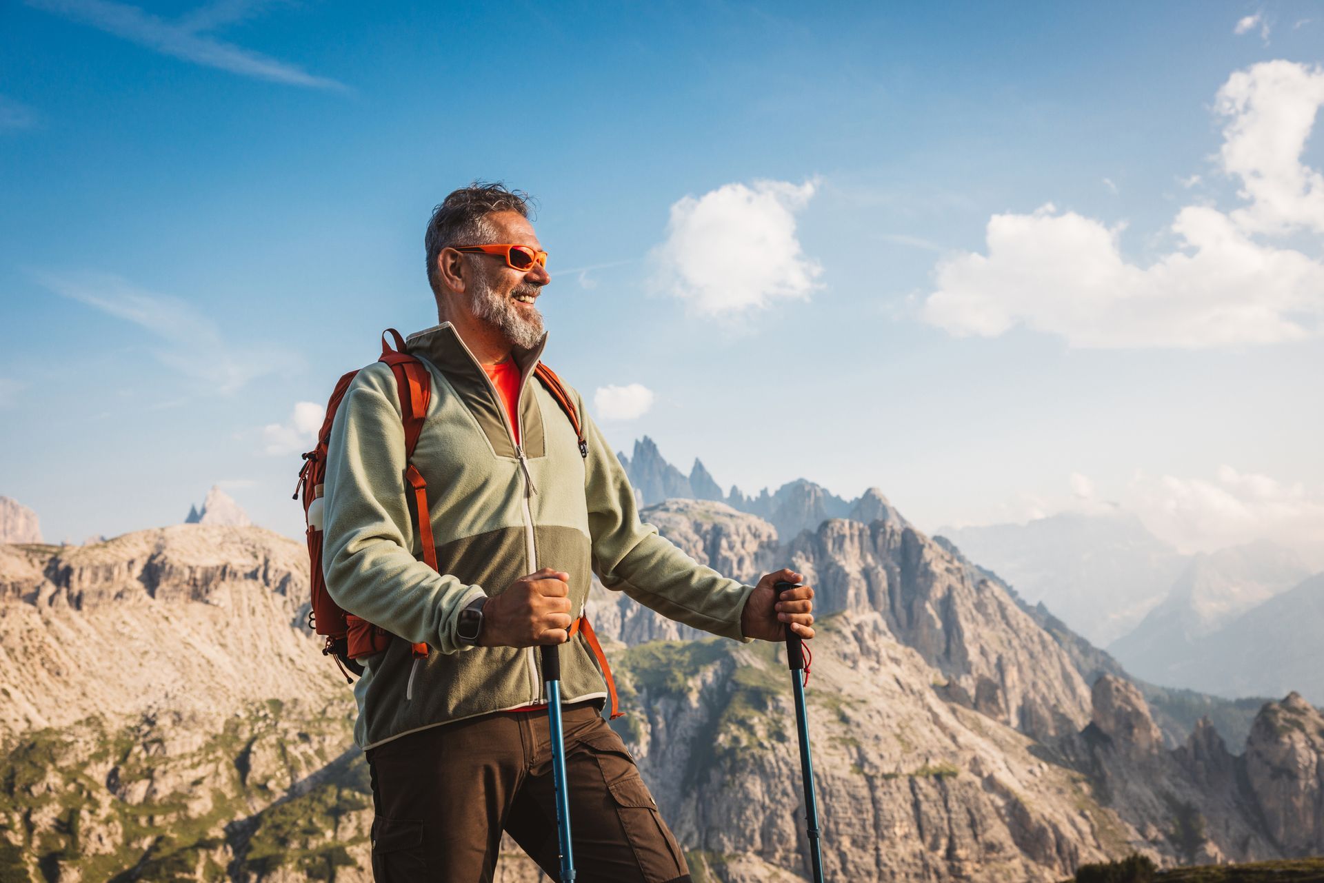 Man hiking in mountains, smiling, using poles, wearing a backpack and sunglasses, under a blue sky.