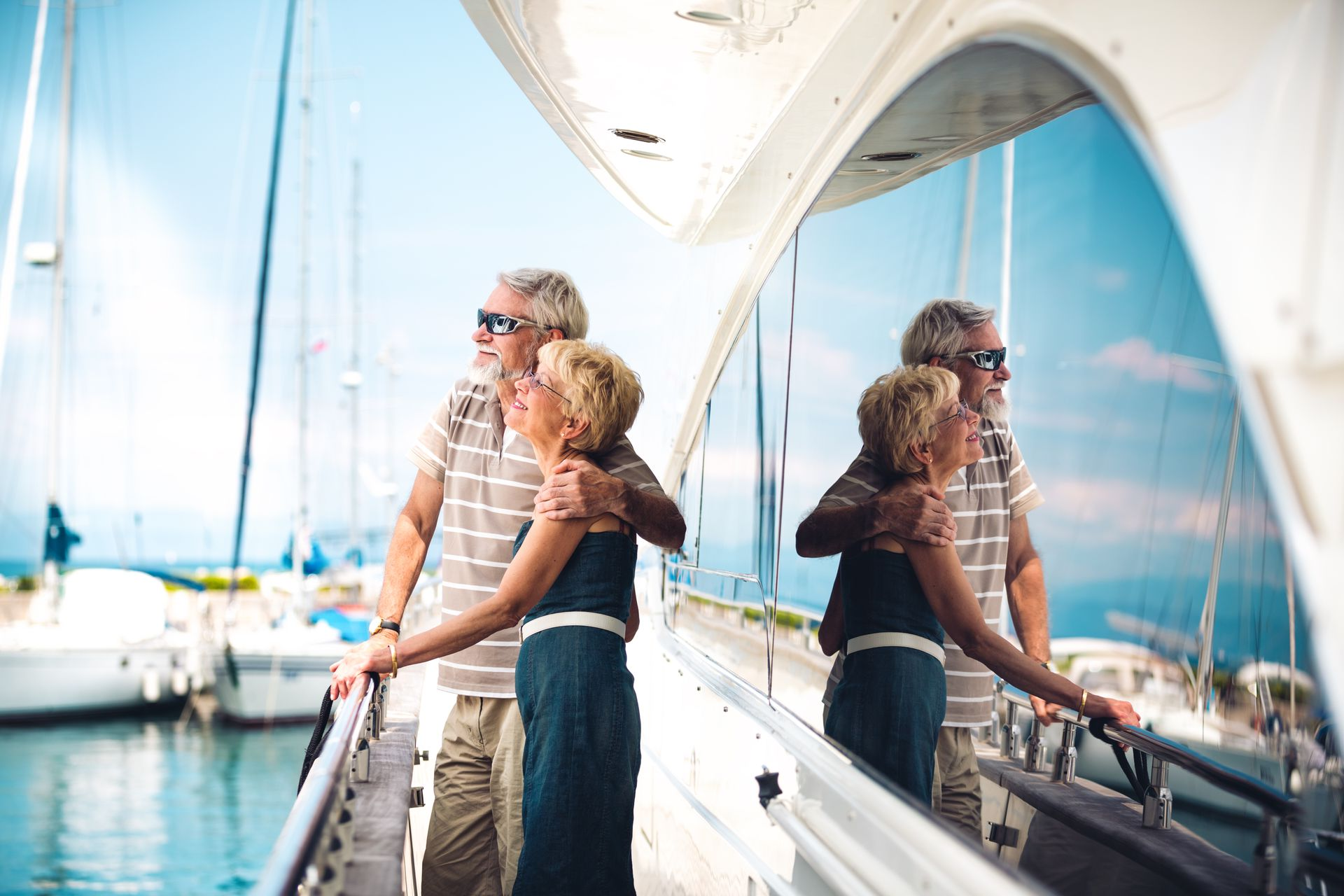 Couple on a yacht overlooking a marina, reflected in glass. The man has his arm around the woman. Blue sky.