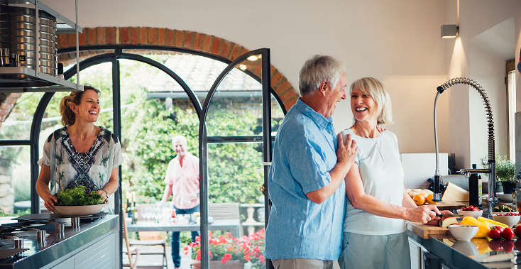 People laughing in a kitchen. Couple dances while others prepare food. Sunlight streams in.
