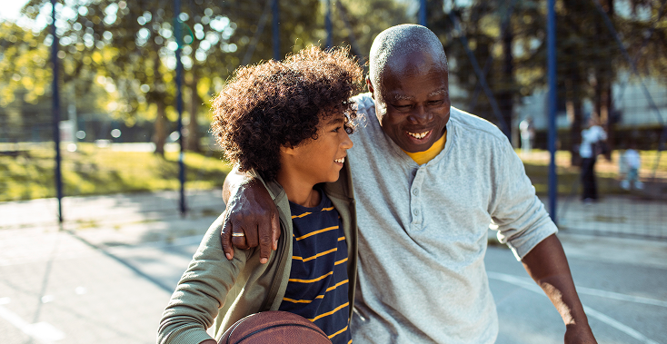 Man with arm around a boy, holding a basketball on an outdoor court, smiling.