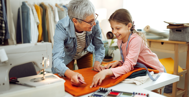 Woman and girl sewing together at a table, smiling. Sewing machine and fabric visible in a well-lit room.