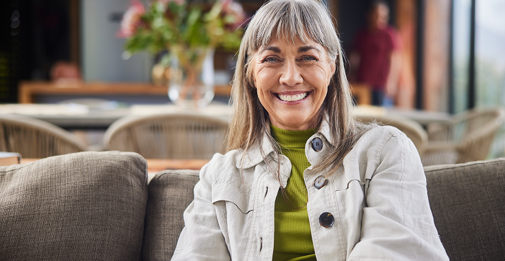 Woman with gray hair smiles, seated on a couch in a well-lit living room, wearing a light jacket over a green turtleneck.