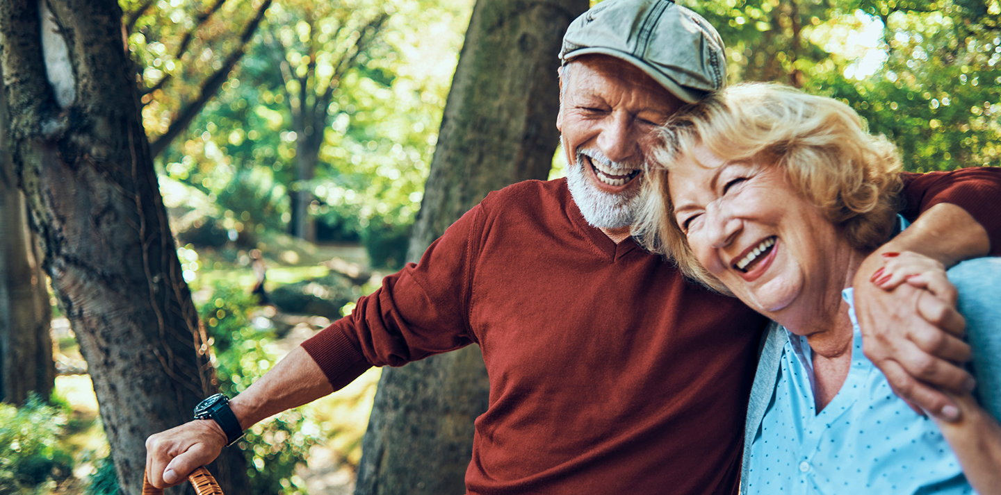 Smiling couple embracing in a forest setting. Man wears a cap and red sweater, woman a light blue top.