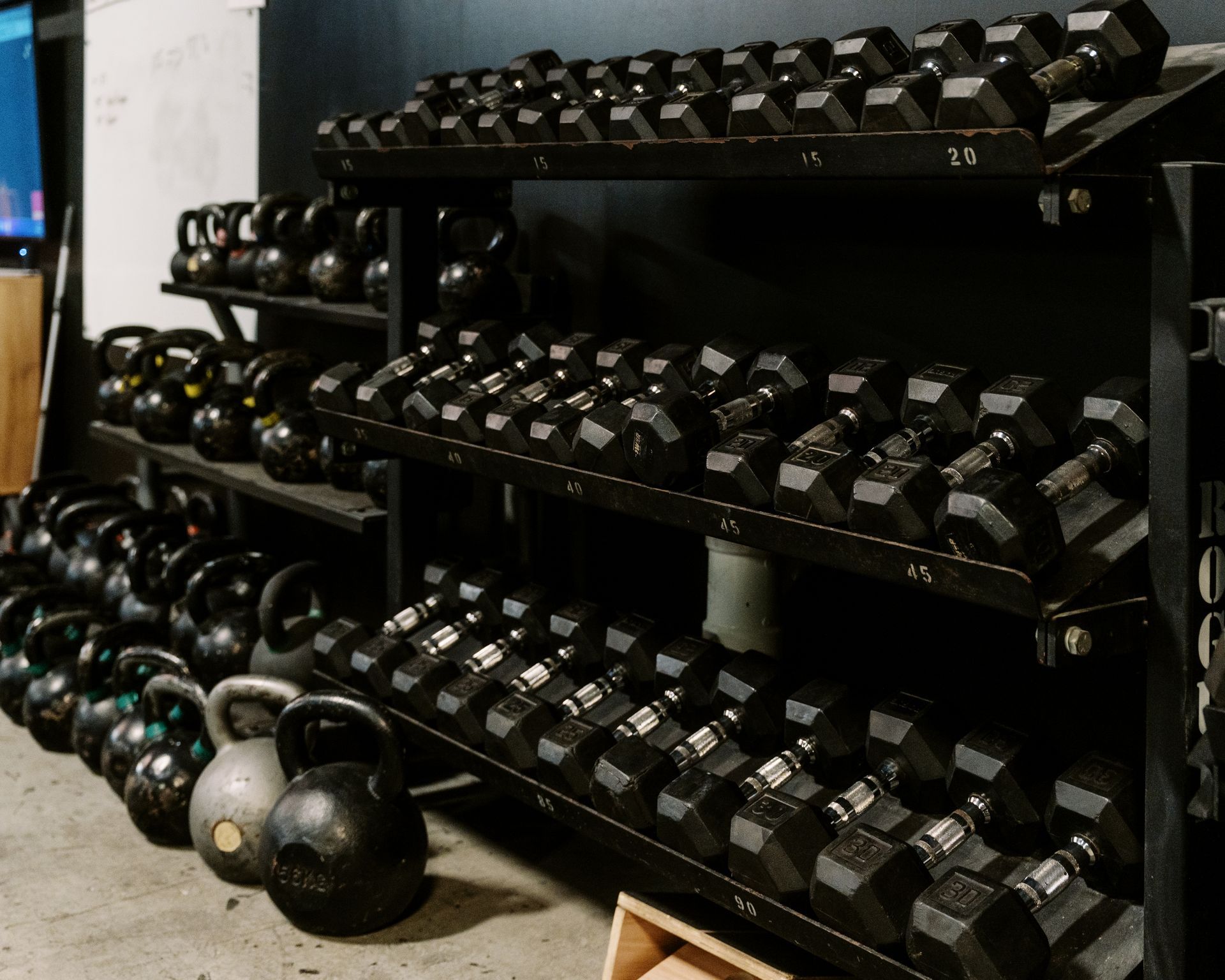 A gym storage rack holding rows of black dumbbells and a collection of kettlebells on the floor.