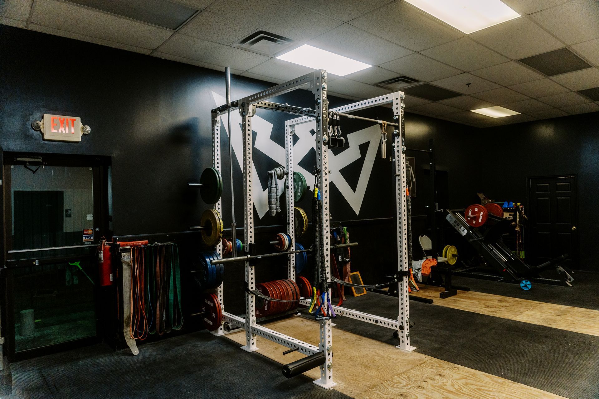 At Be Somebody Gym in Knoxville, TN, the interior features a white squat rack, weight plates, and resistance bands against a black wall with a white logo.