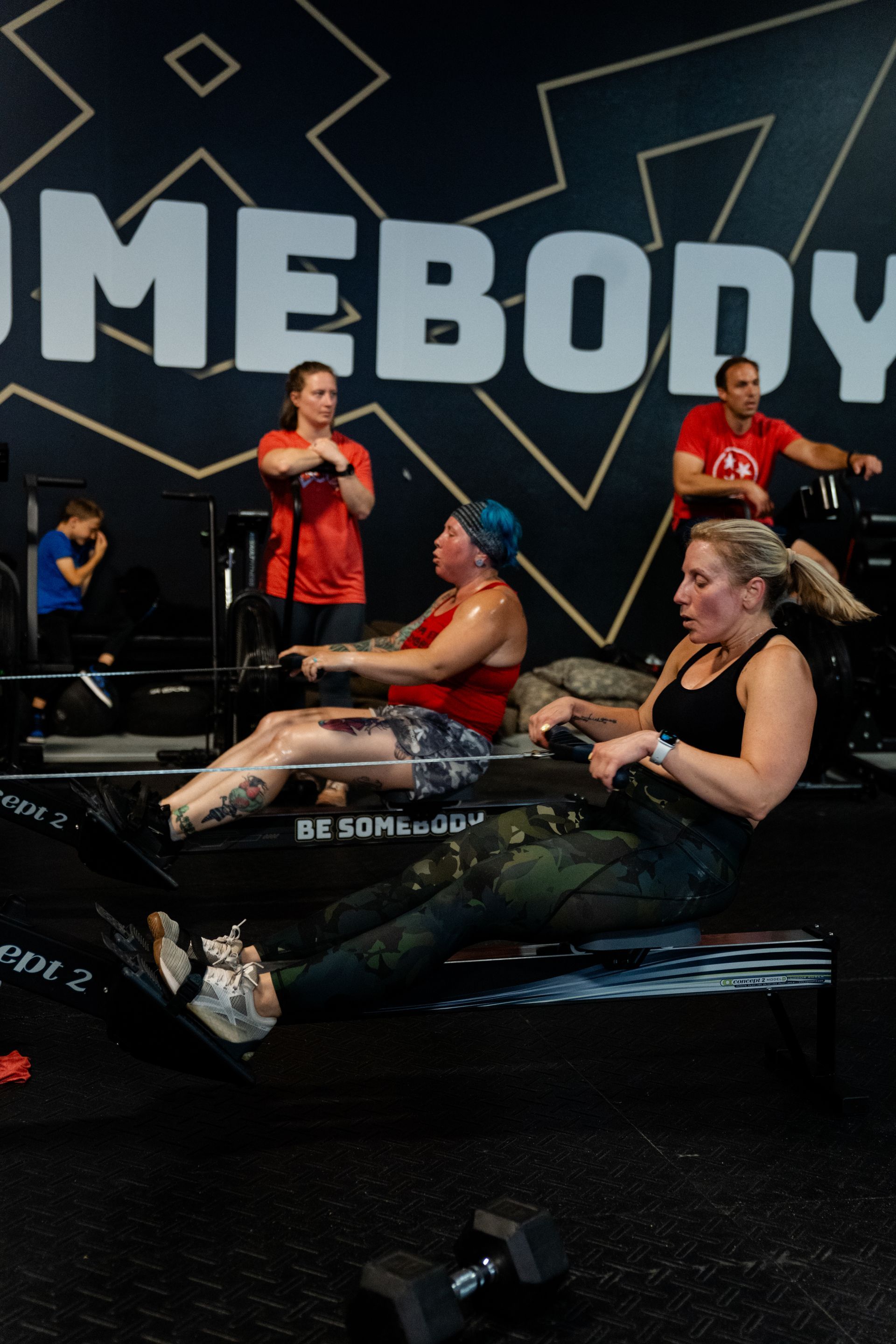 Three people use rowing machines at Be Somebody Gym in Knoxville, TN with a black and gold wall, with a weight on the floor in the foreground.