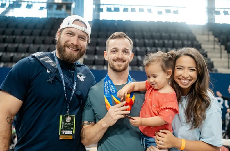 Three people pose smiling in an arena, with a man in the center holding a medal and a child in his arms.