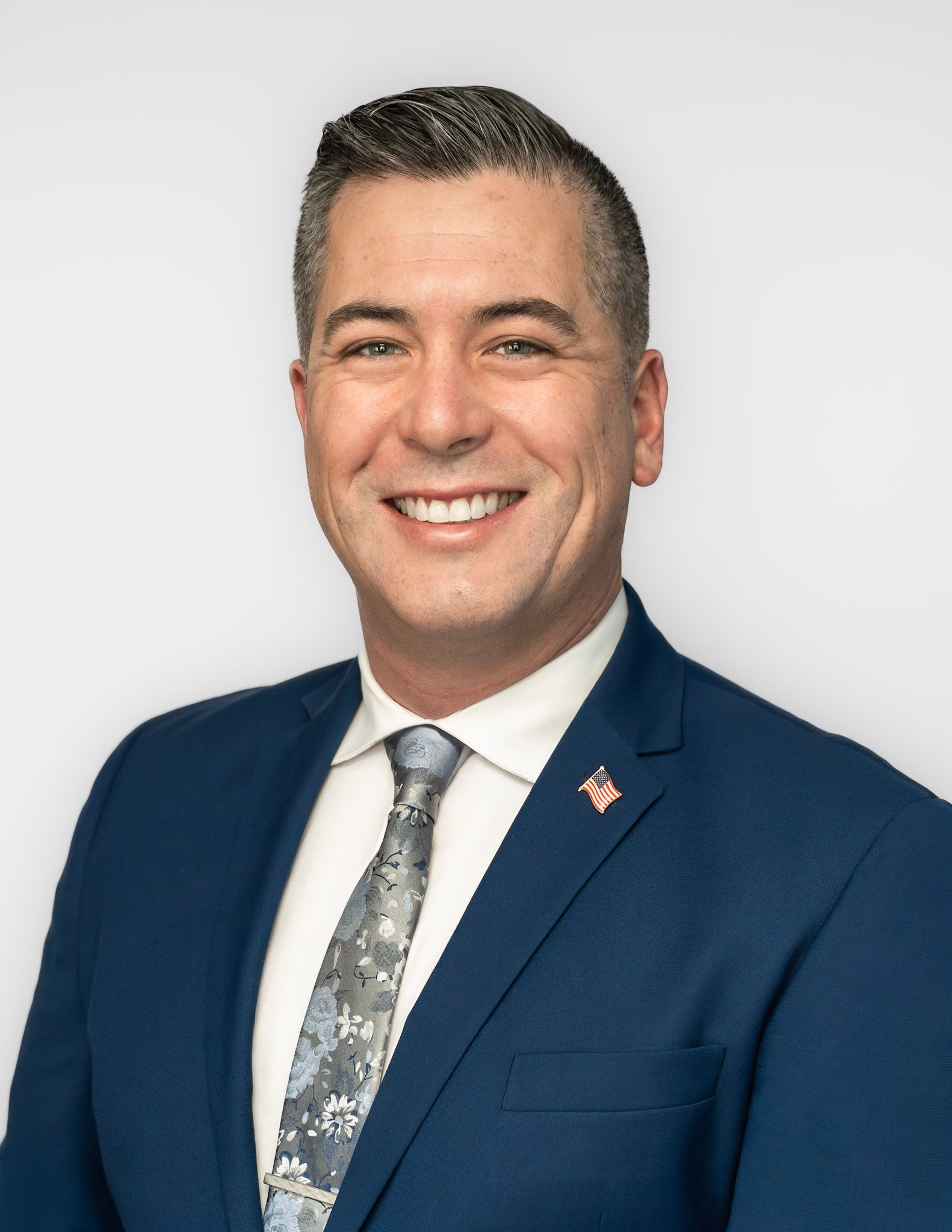 Man in a blue suit smiles, wearing a patterned tie, against a white background.