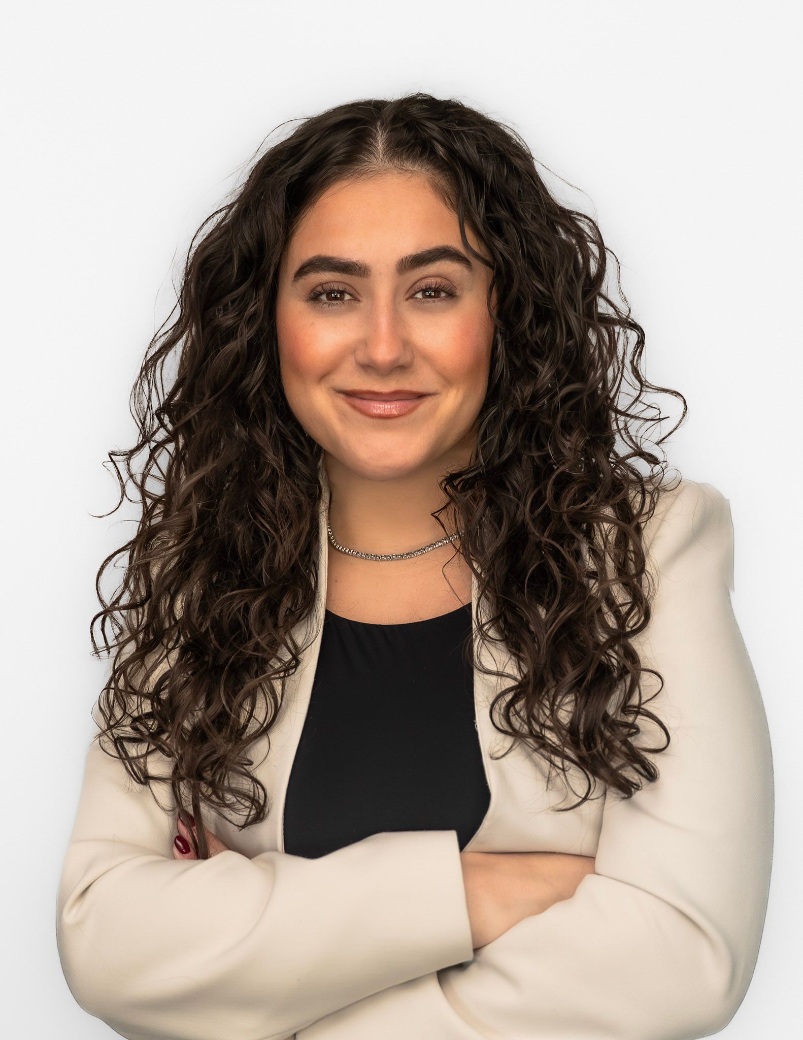 Woman with curly hair, arms crossed, wearing a beige blazer and black top, smiling against a white background.