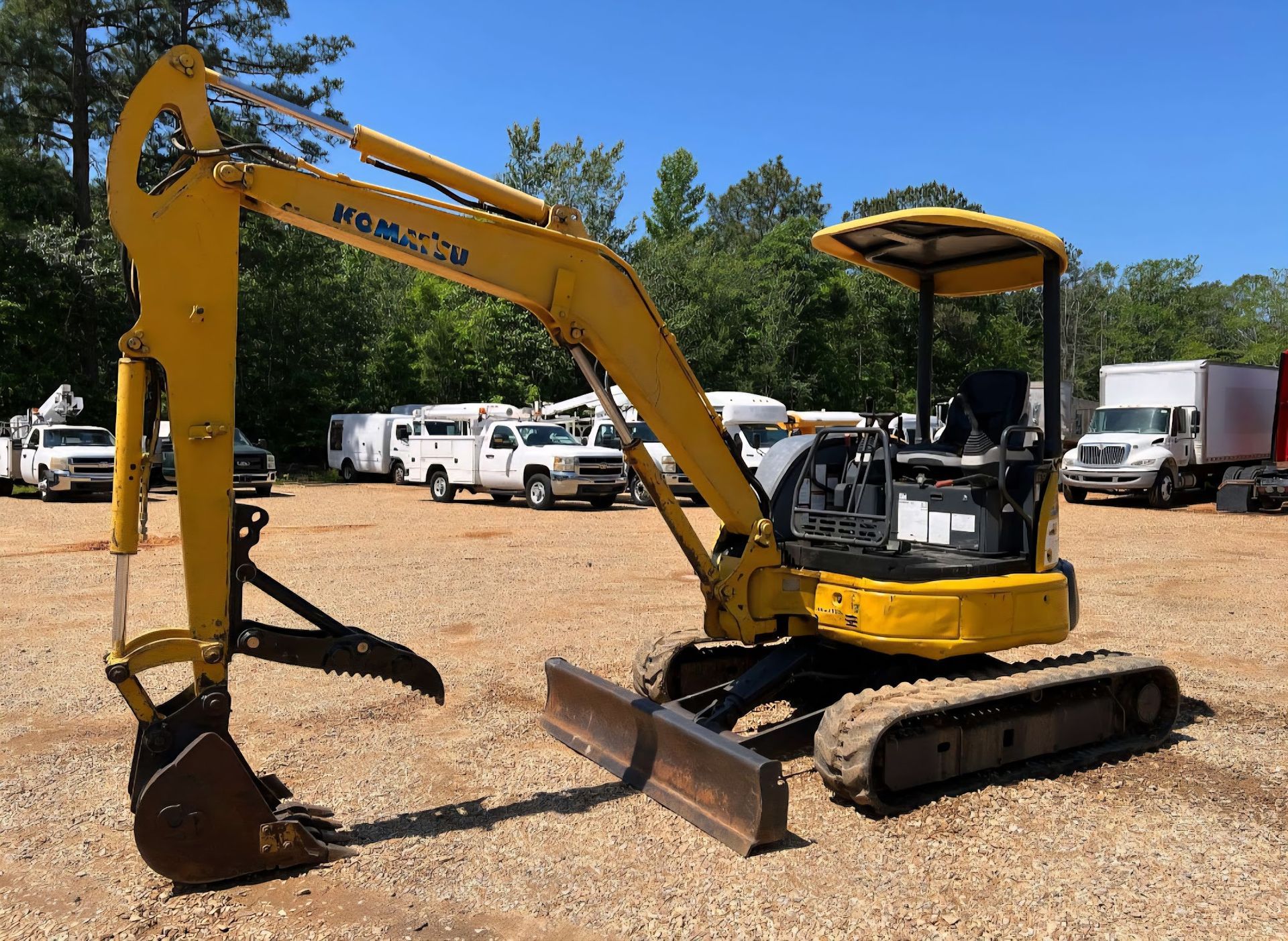 Yellow Komatsu mini excavator on gravel, parked outdoors with trucks and trees in the background.