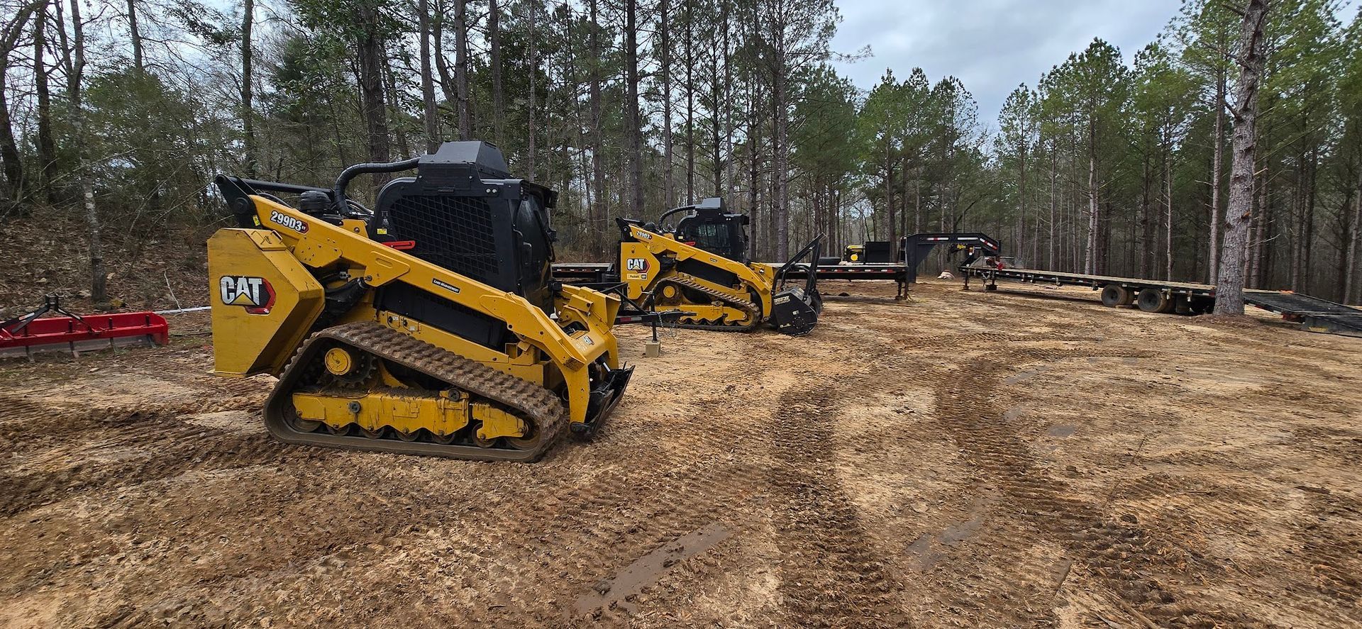 Yellow CAT skid steers working in a dirt clearing, surrounded by trees.