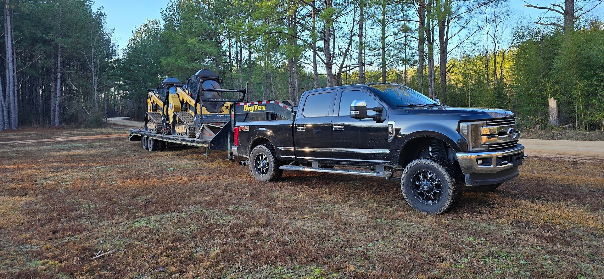 A black truck towing a trailer with a piece of heavy construction equipment on a dirt road, trees in the background.