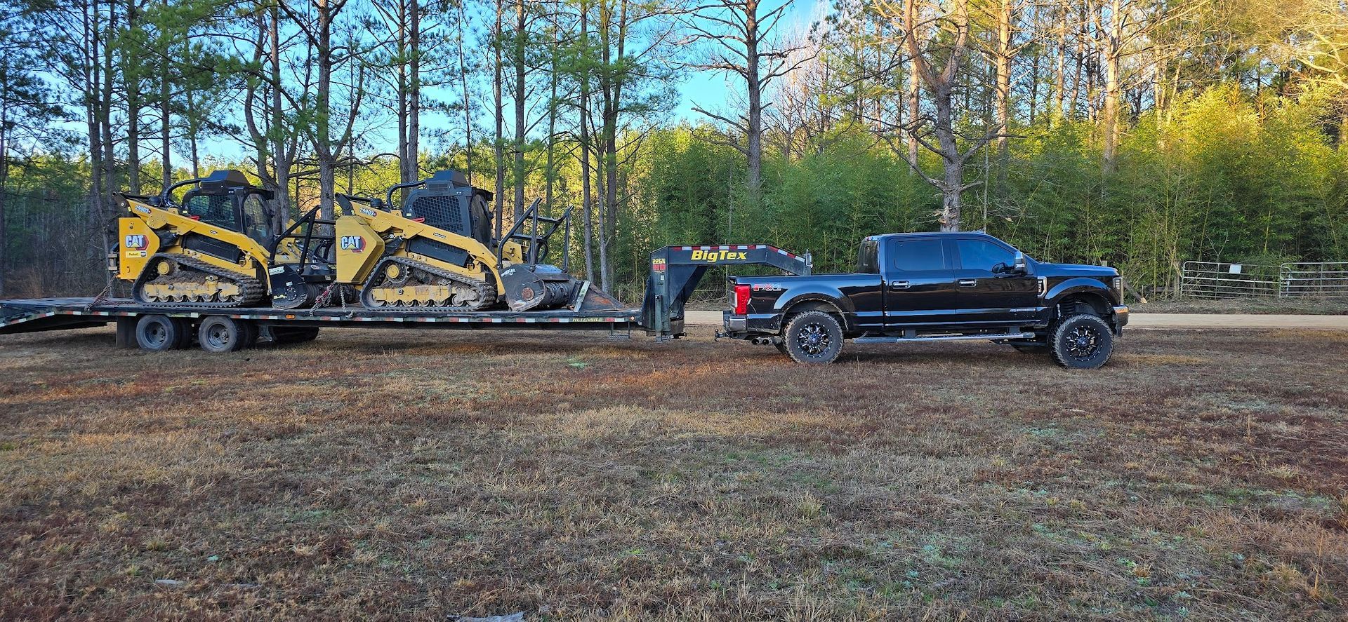 Black truck towing a trailer carrying yellow construction equipment on a dirt road in front of trees.