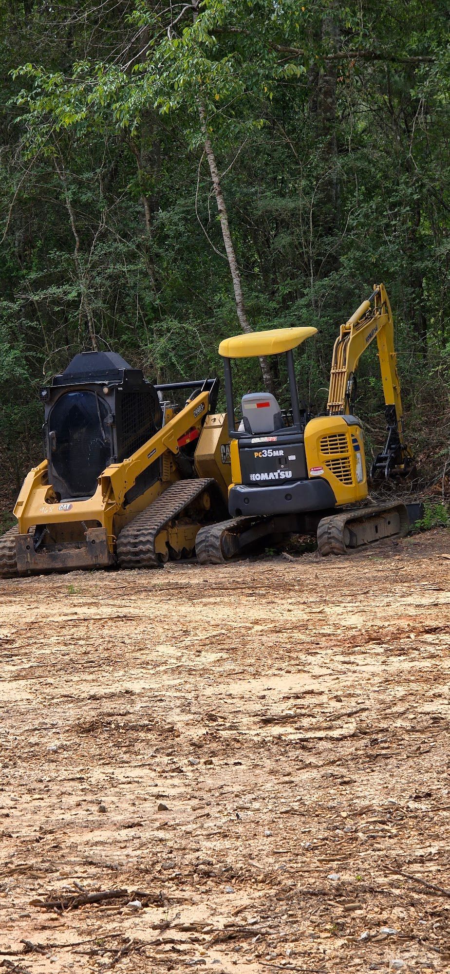 Two yellow construction vehicles on a cleared dirt area with trees in the background.