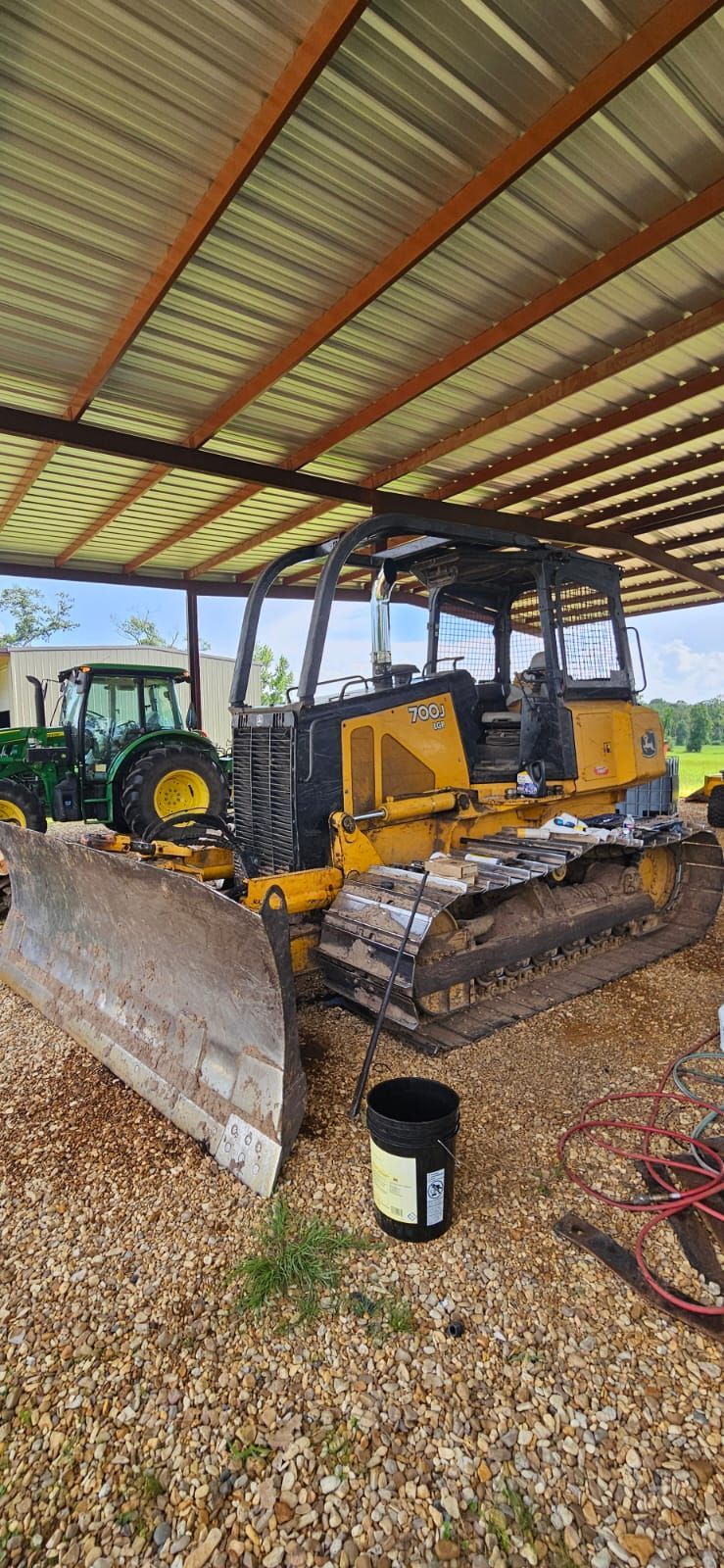 Yellow bulldozer under a metal roof, parked on gravel. A green tractor is visible in the background.