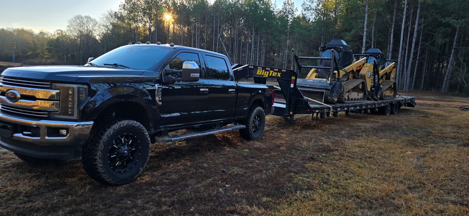Black truck pulling a trailer with yellow construction equipment parked in a wooded area.