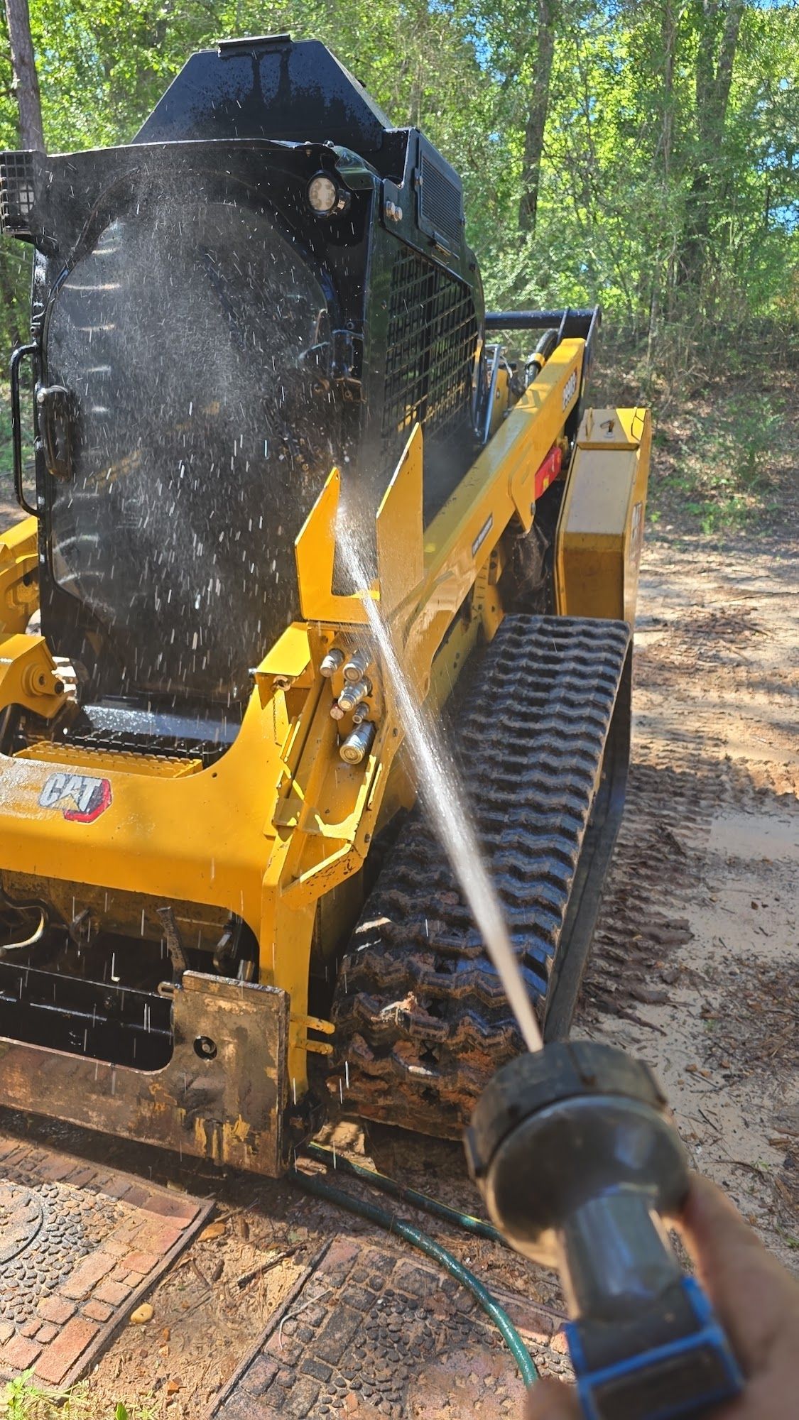 A person sprays water on a yellow and black skid steer loader. The loader has tracks and is outdoors.