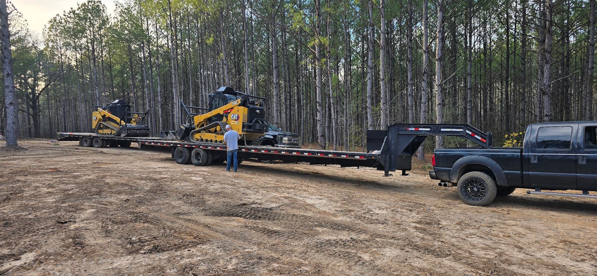 A truck pulling a trailer with two yellow construction vehicles in a wooded area. Man stands beside.