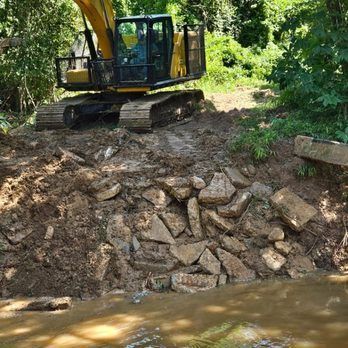 Yellow excavator on muddy bank, moving broken concrete blocks near a stream.