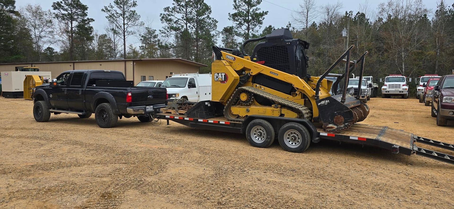 A black truck towing a yellow skid steer loader on a trailer in a gravel lot.