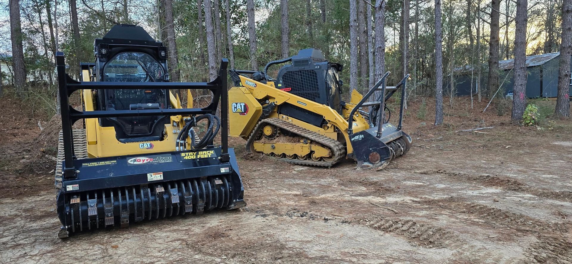 Two yellow skid steers with attachments sit in a wooded area.