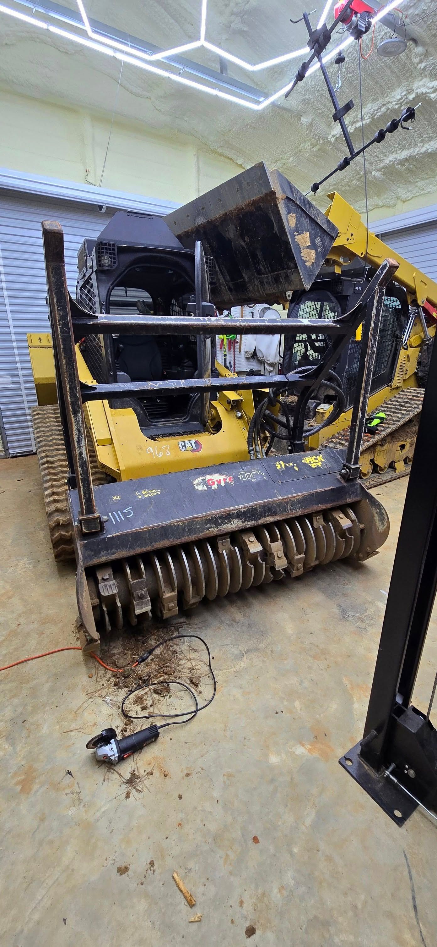 Yellow and black forestry mulcher machine inside a garage, with overhead lights and a debris covered floor.