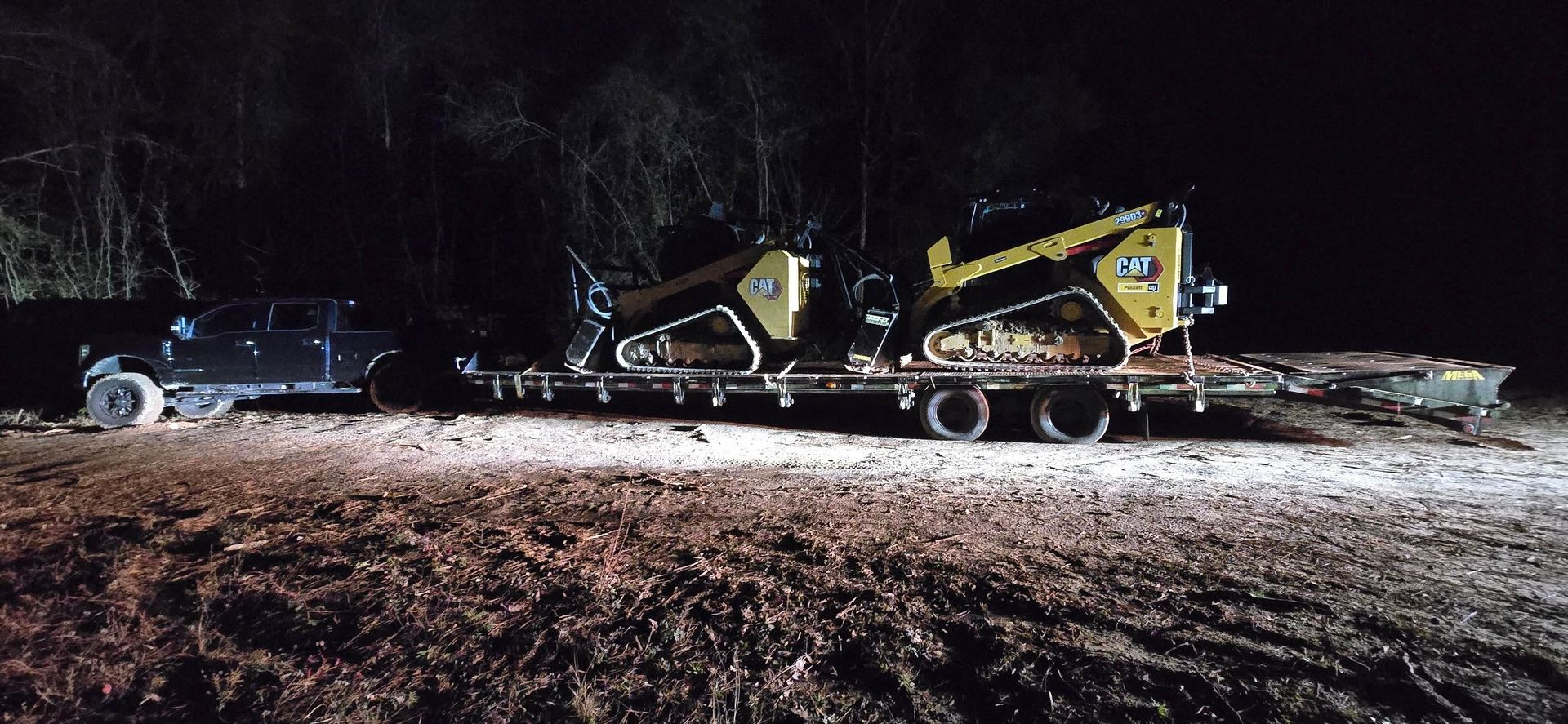A black truck pulling a flatbed trailer carrying two yellow and black construction machines at night.