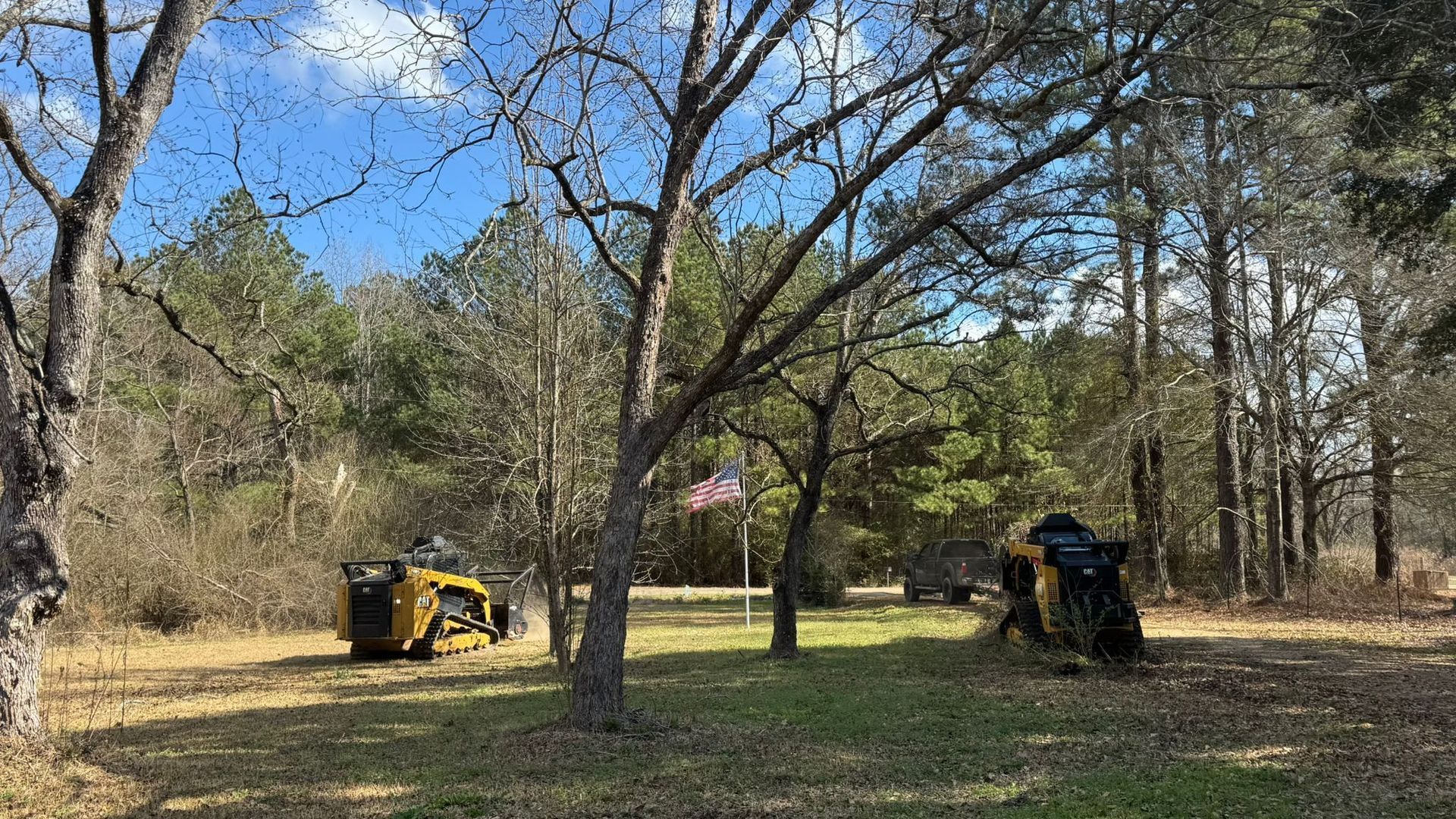 Two yellow tractors clearing land near trees, with an American flag in the background.