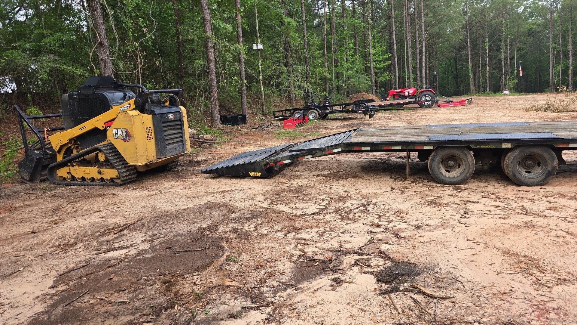 Yellow skid steer loader on muddy ground near a flatbed trailer in a wooded area.