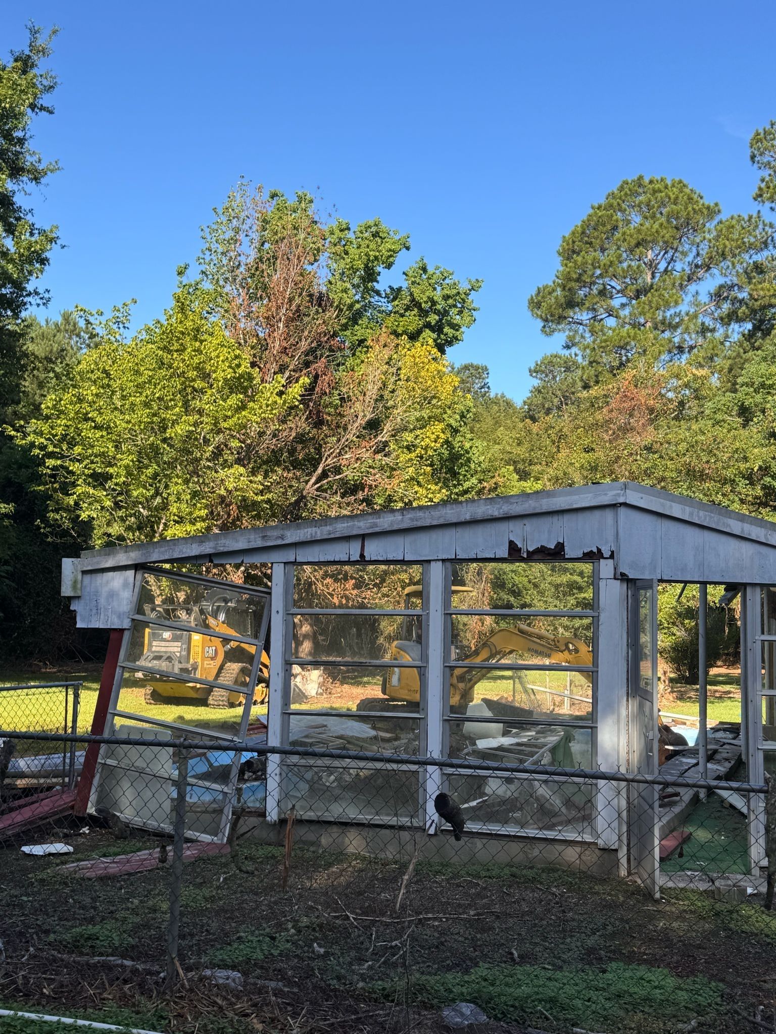 A weathered outdoor enclosure with glass panels, surrounded by trees under a blue sky.