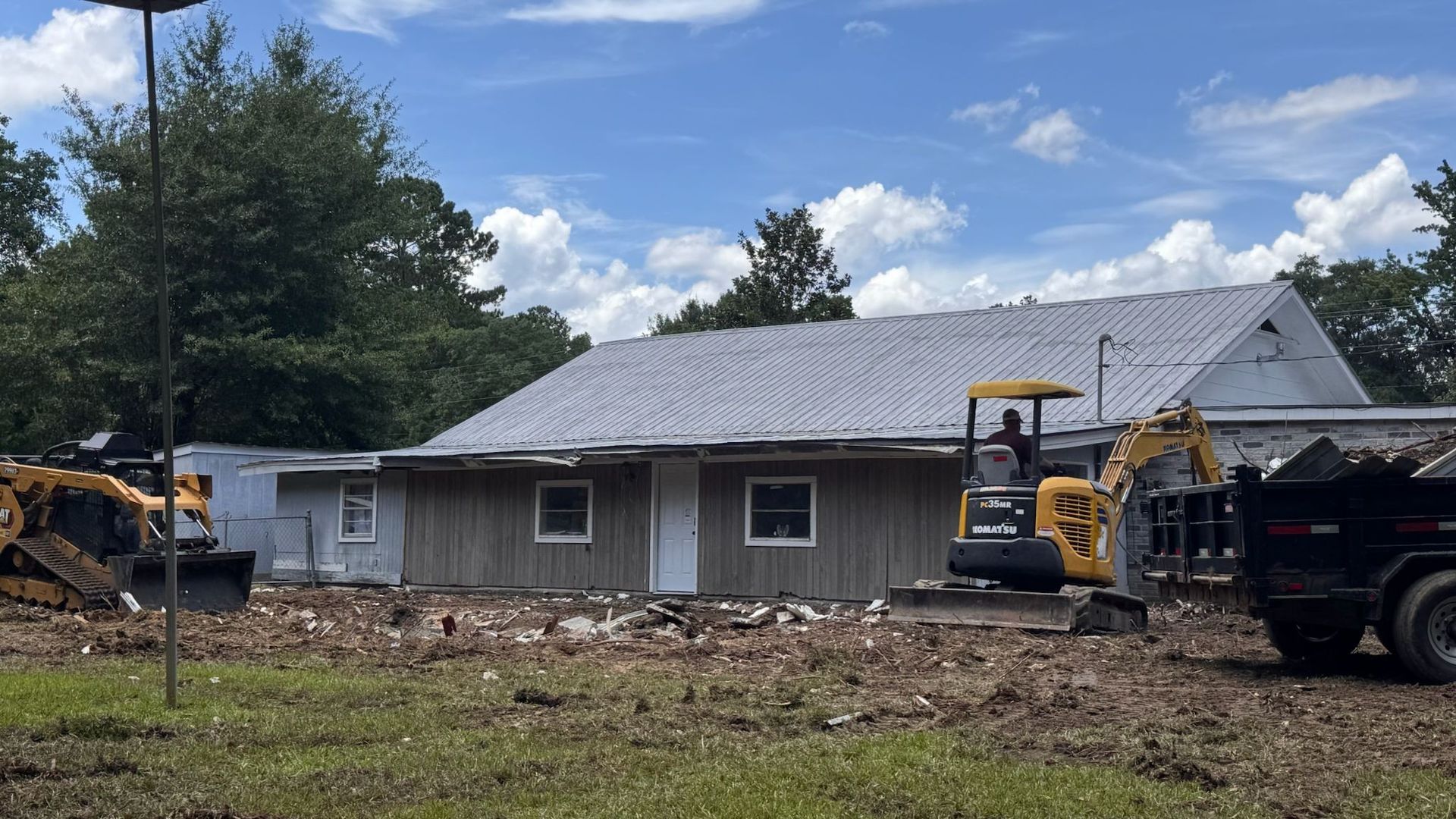 Construction site with a building, excavator, and dump truck on a brown field under a blue sky.