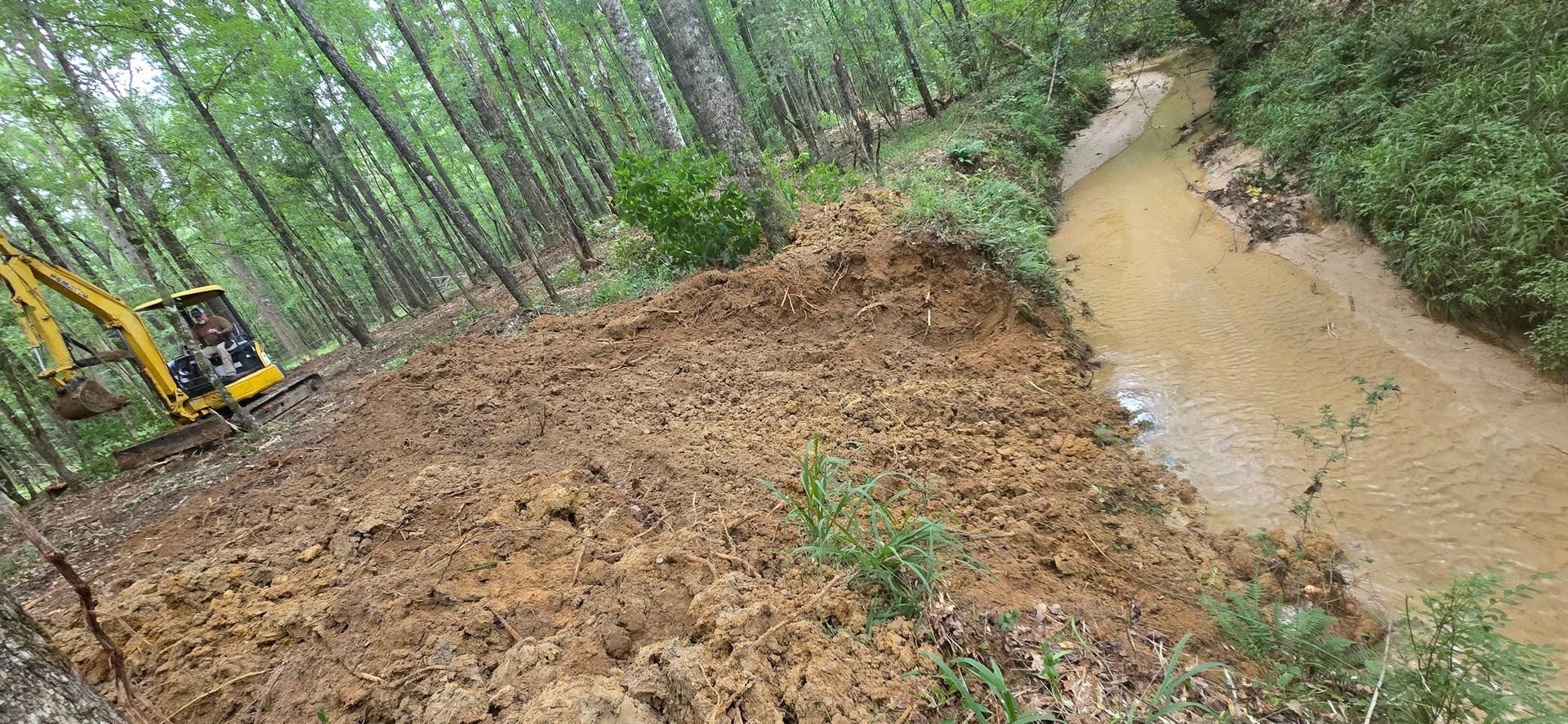 A yellow excavator near a muddy embankment in a forest beside a brown water stream.