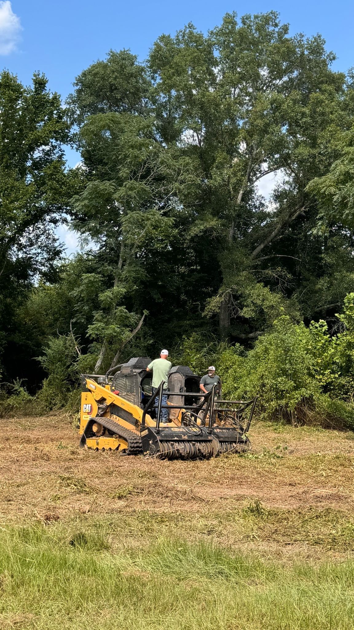 Two people operate a yellow skid steer with a mulching head, clearing a field near trees on a sunny day.