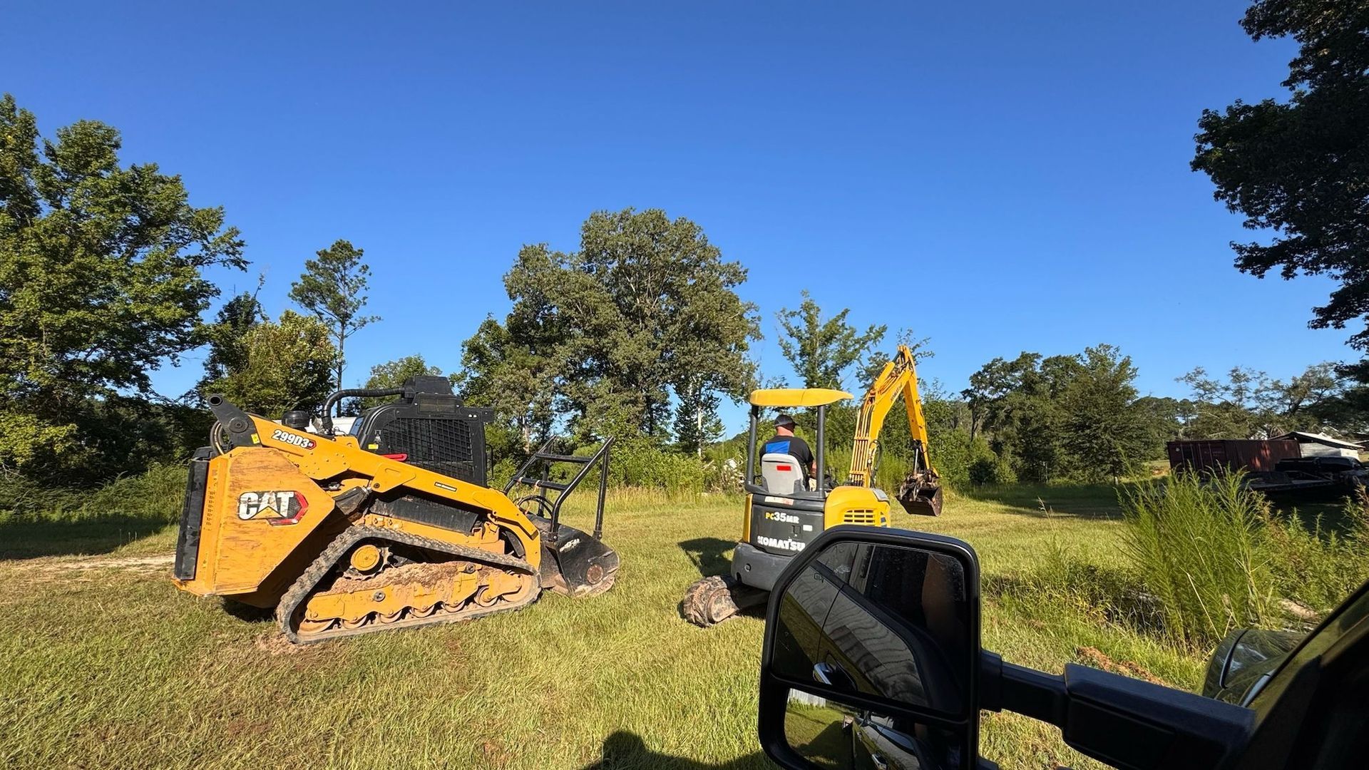 Yellow construction equipment in a grassy field on a sunny day. A person operates a backhoe.