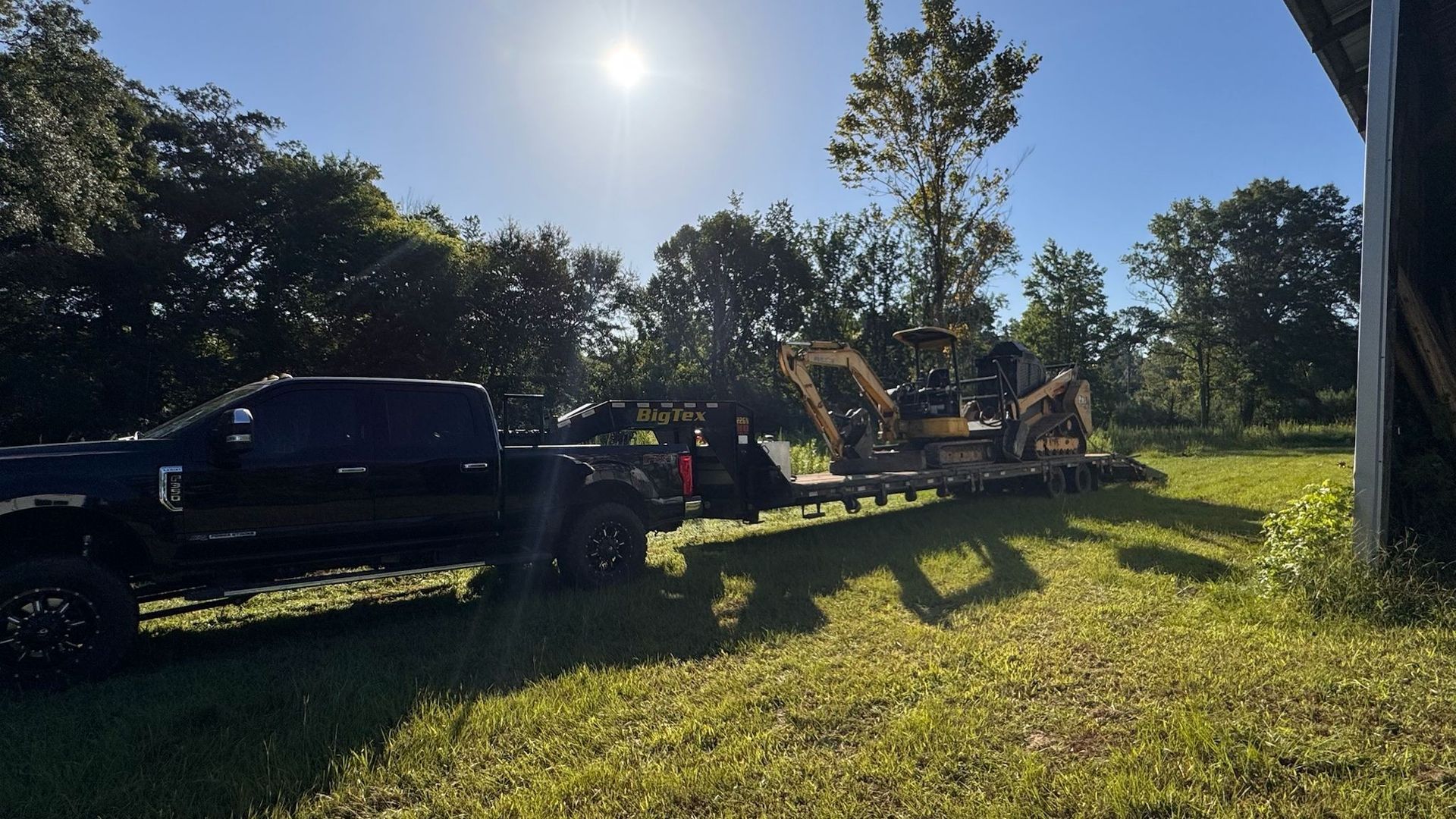 Black truck towing a trailer with two yellow excavators on a grassy field; sunny day.