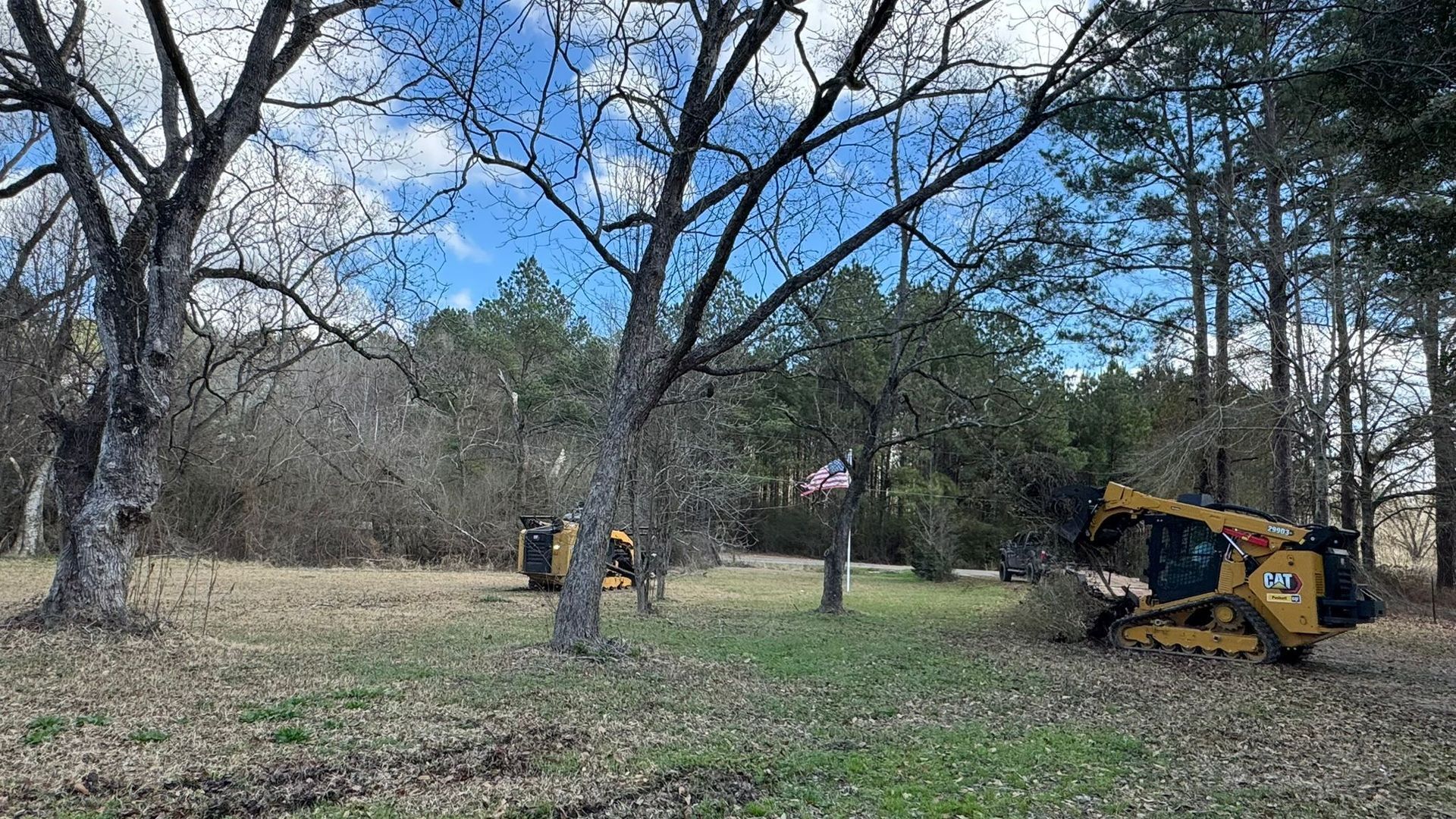 Two yellow construction vehicles clearing a grassy field with trees under a blue sky.