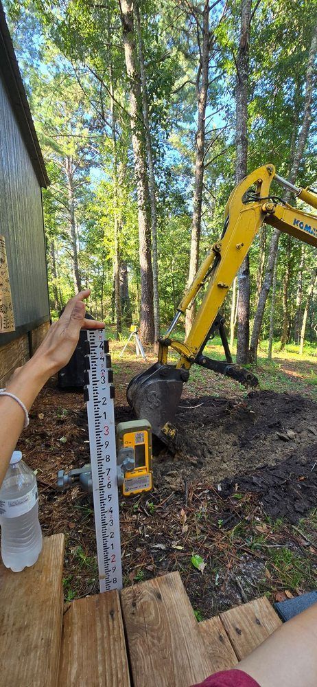 A yellow excavator digging in a yard; a person measures the depth with a ruler.