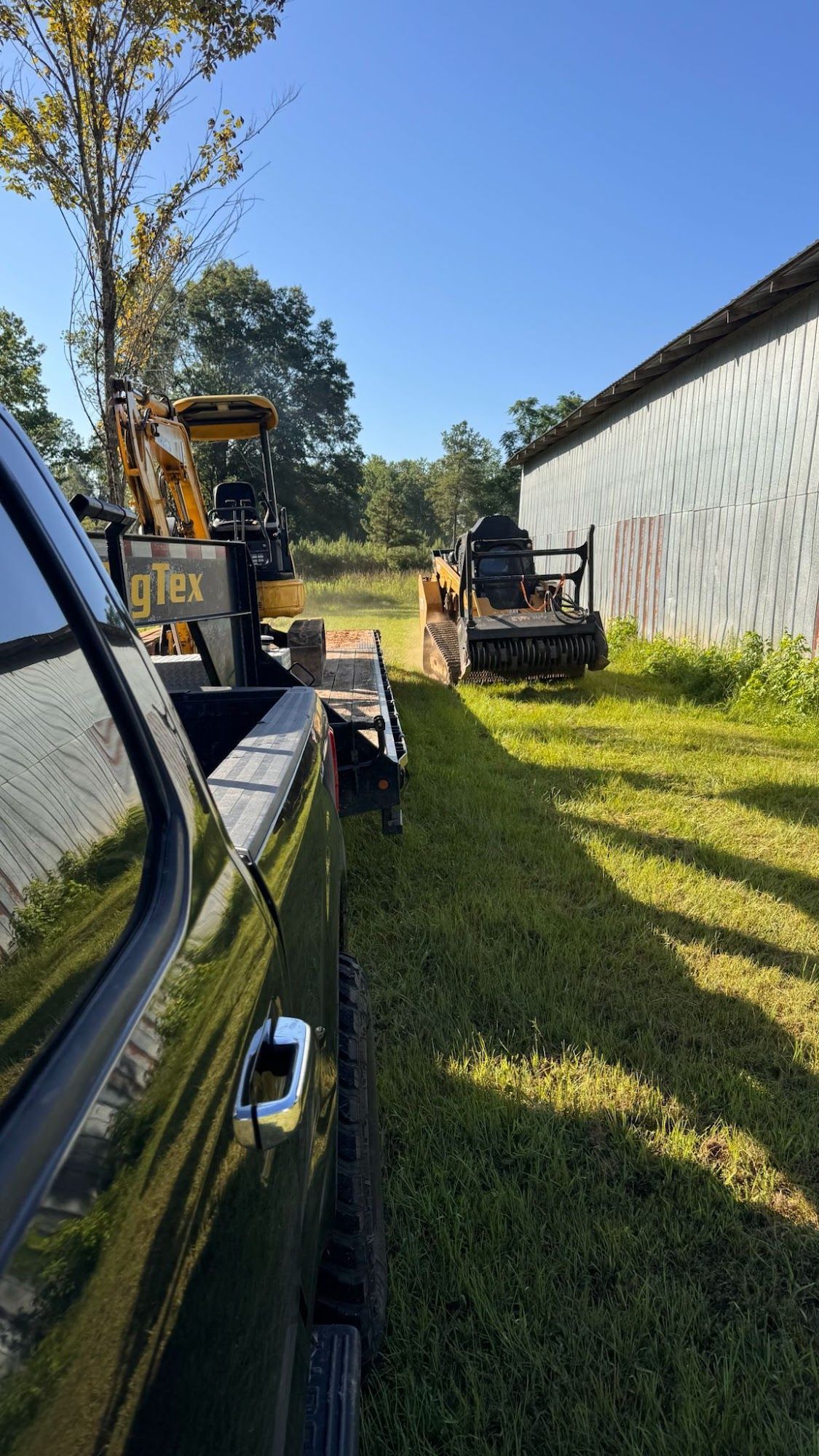 Truck with trailer holding machinery near a barn on a sunny day.