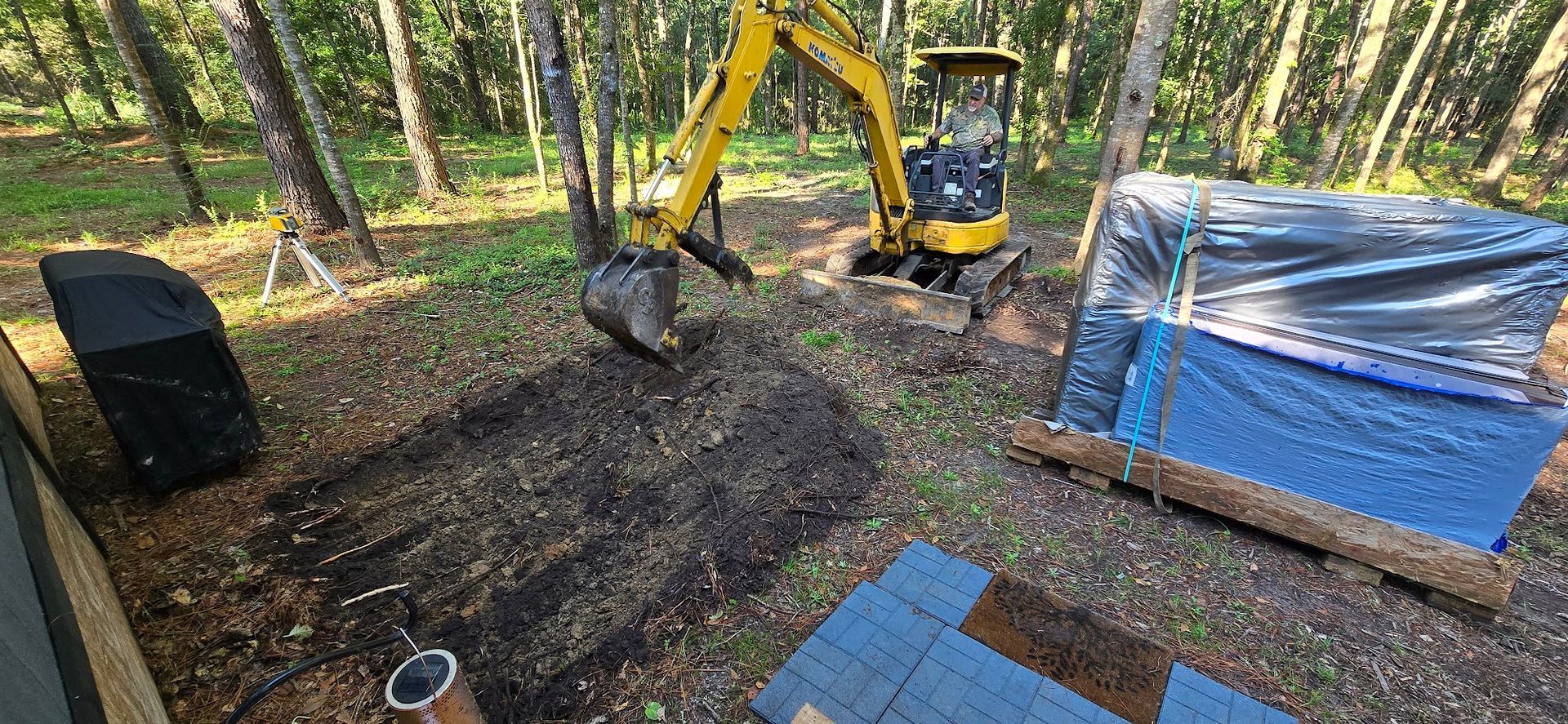 Yellow excavator digging in a wooded area; blue tarp-covered item on a wooden pallet nearby.