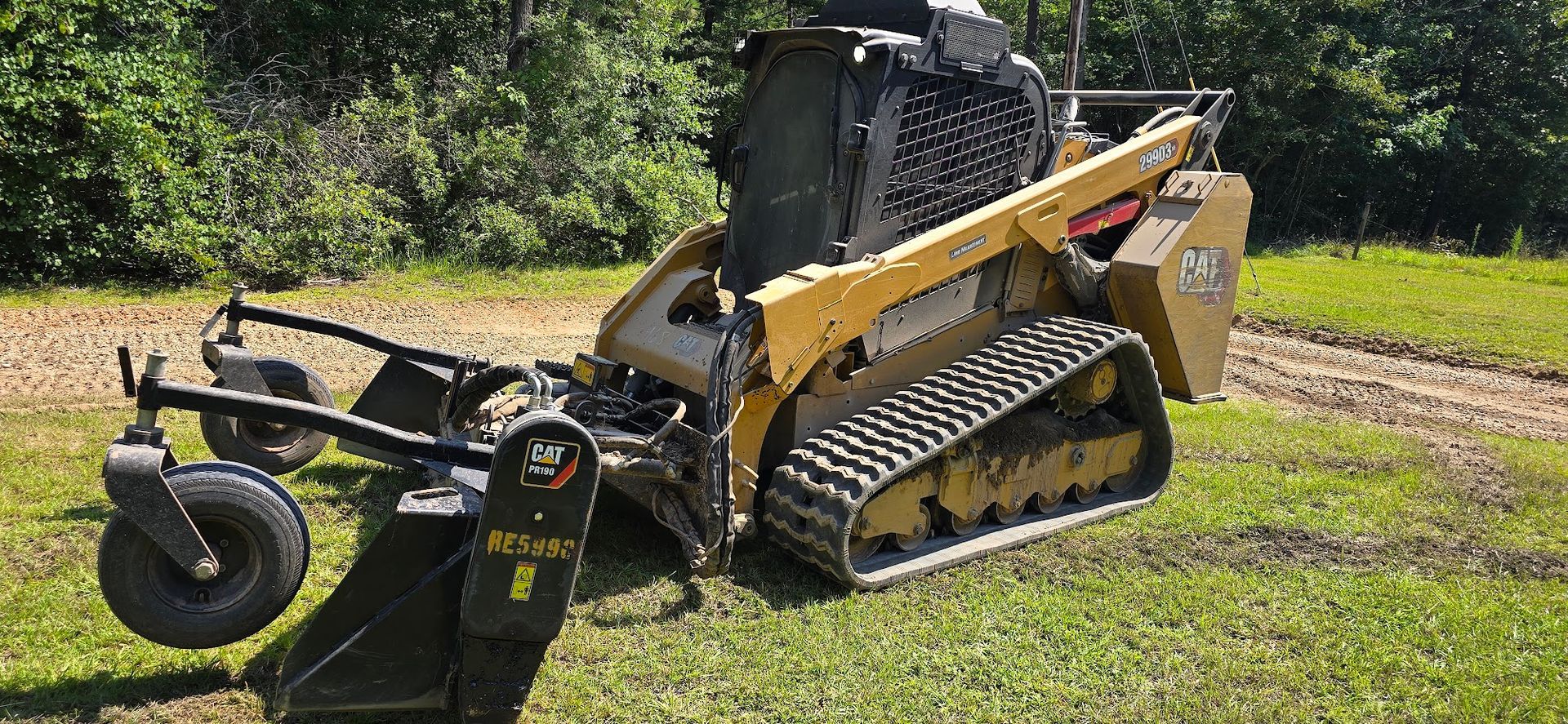 Yellow track skid steer with brush cutter attachment on grass.