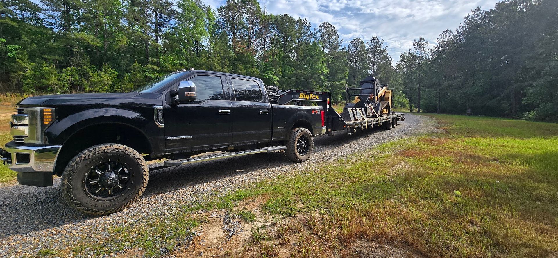 Black truck hauling a trailer with construction equipment on a gravel road, surrounded by trees.