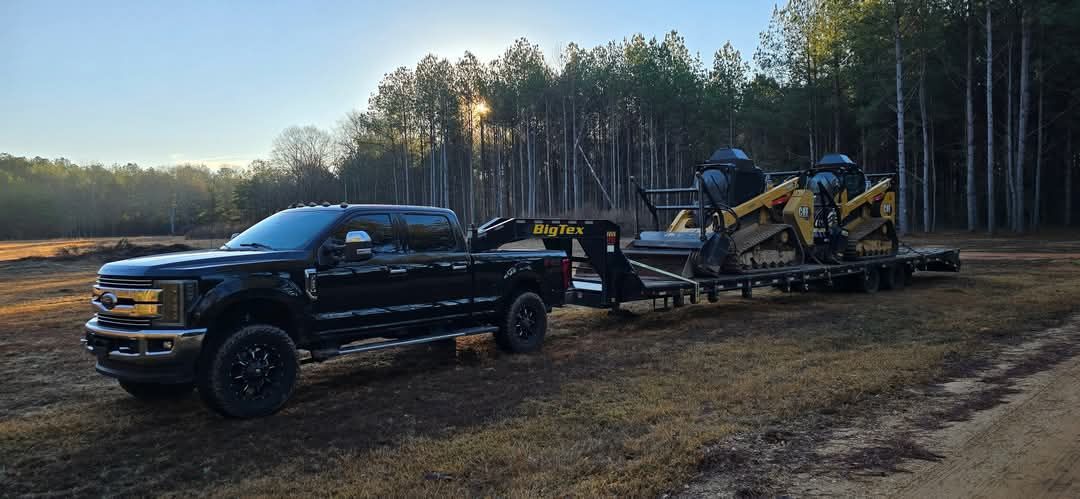 Black pickup truck towing machinery on a trailer, in a field, with forest background.