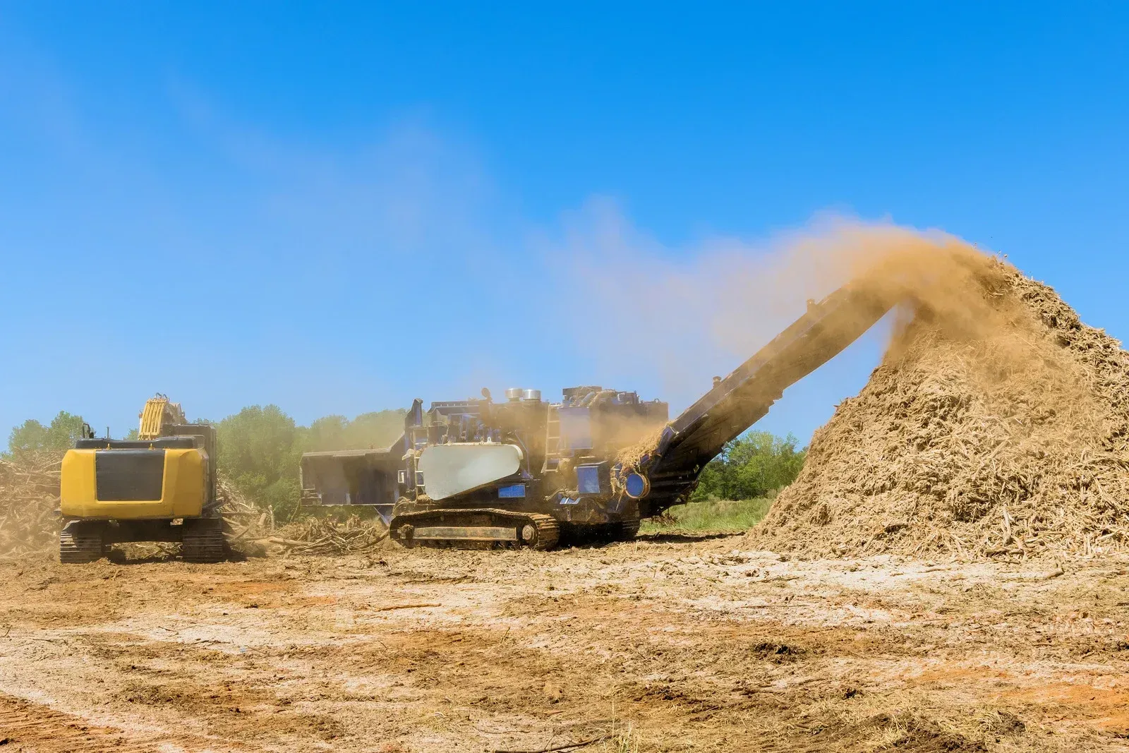 Construction site with a building, excavator, and dump truck on a brown field under a blue sky.