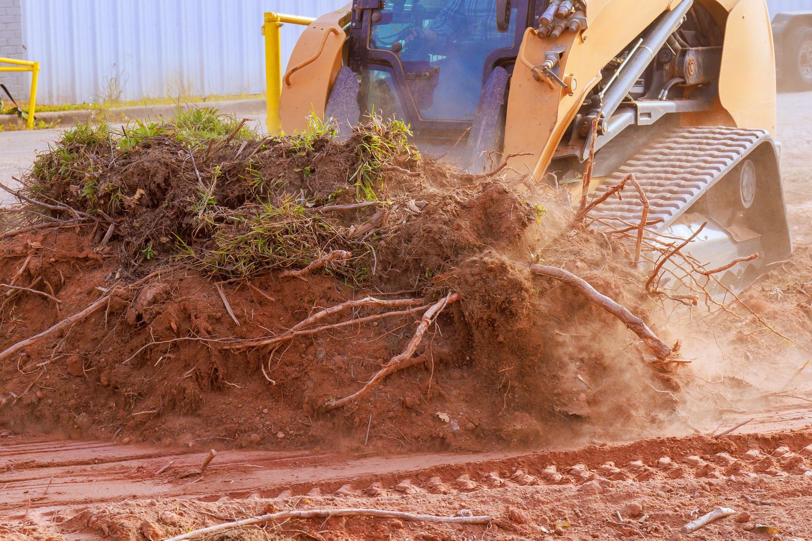 A yellow excavator works on muddy terrain beside a stream in a forest. Green trees line the banks.