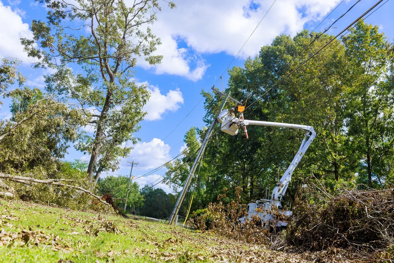 Utility worker in high-visibility vest repairs power lines from a bucket truck after a storm. Blue sky, green trees.
