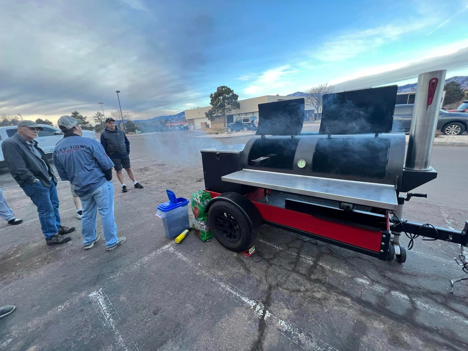 A group of men are standing around a large grill on a trailer in a parking lot.